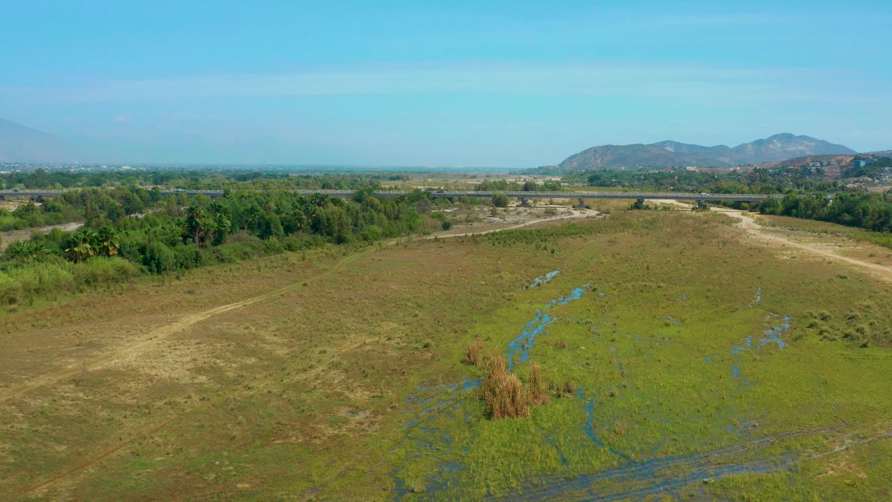 Aerial view over an empty green pasture in San Jose Del Cabo, Mexico, with blue water channels, tree lines, distant mountains, and no visible people.