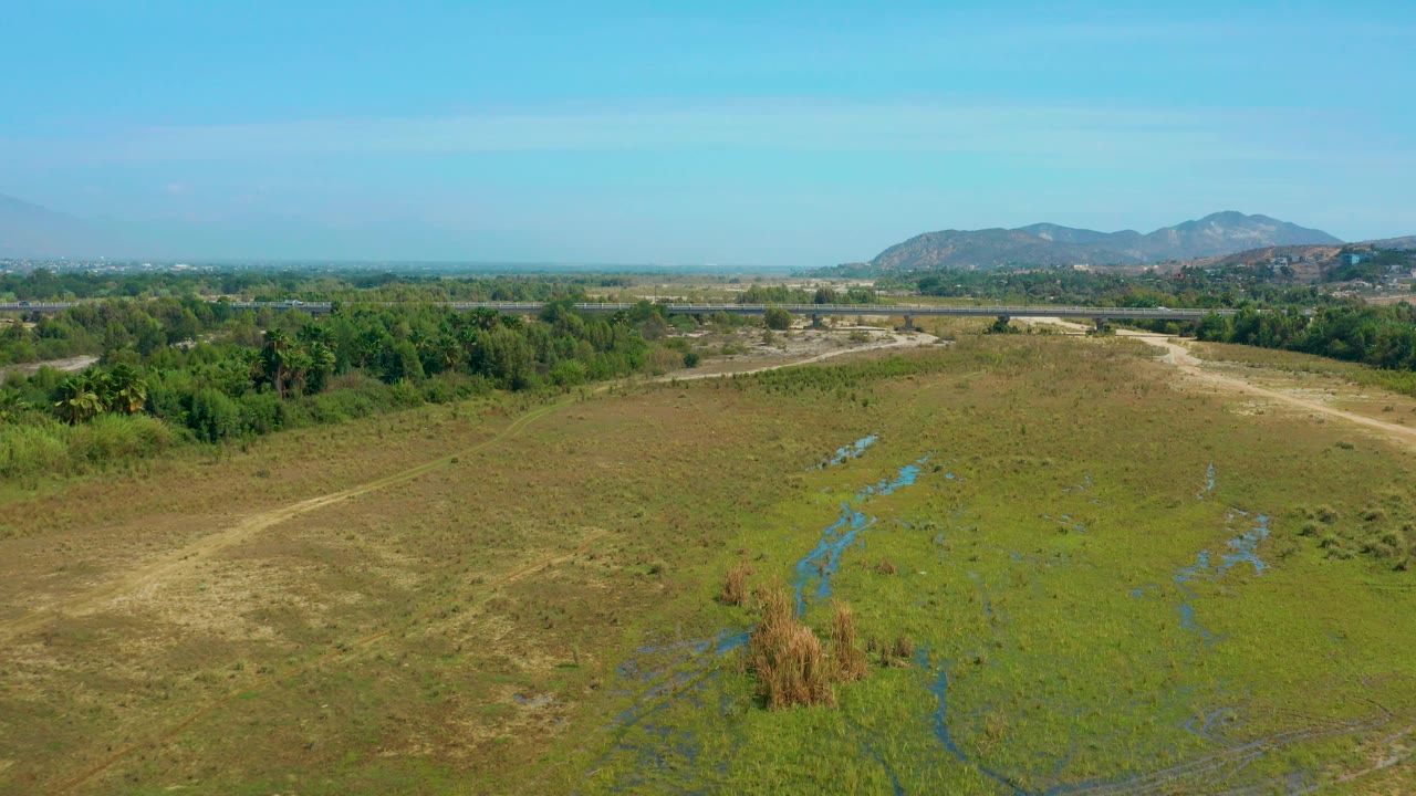 High aerial view in San Jose Del Cabo, Mexico, with an empty green field, small reed clusters, blue water channels, dense trees, low mountains, and no visible people.