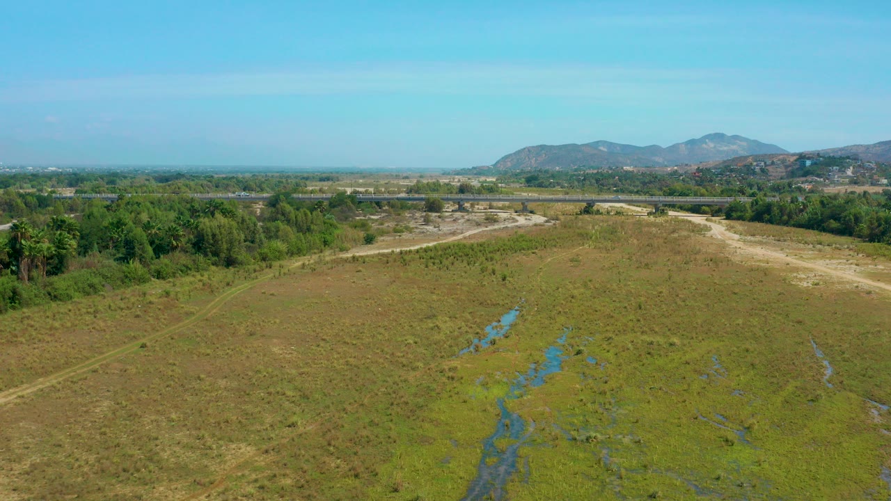 Opening aerial frame over San Jose Del Cabo countryside with wet channels in a green-brown field, tree line, distant mountains, blue sky, and no visible people.