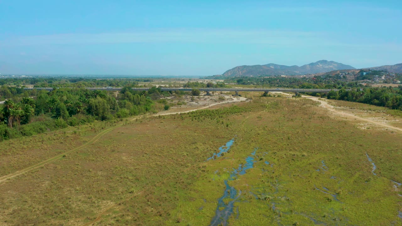 San Jose Del Cabo aerial view one second in, with reflective wet strips cutting through the field, pale dirt paths at the edges, tree cover beyond, and no visible people.