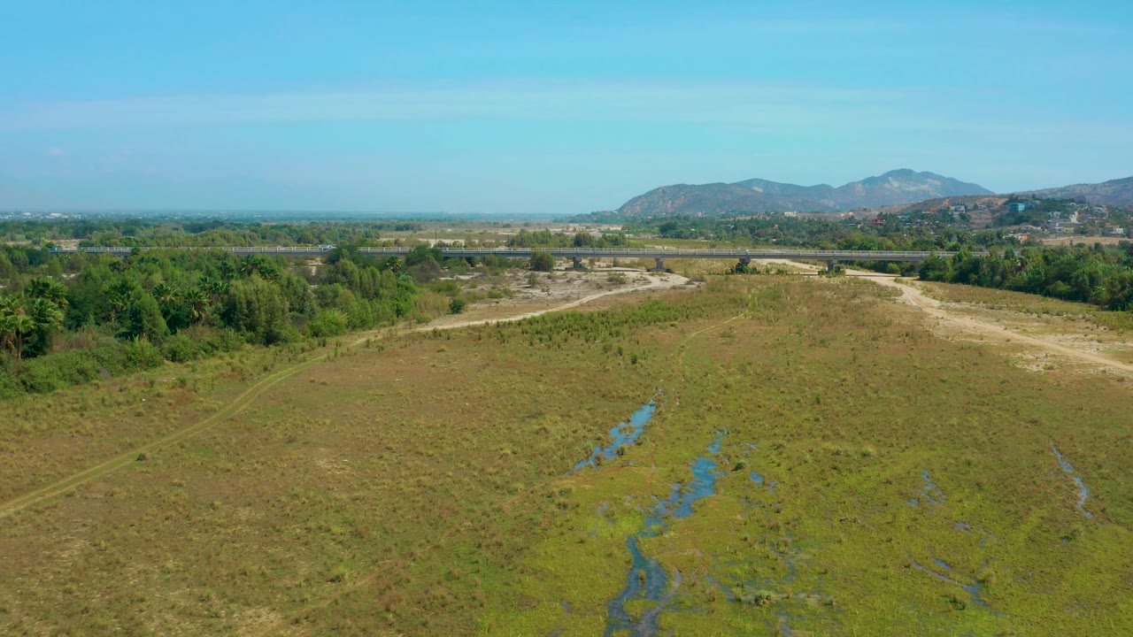 A high drone frame over San Jose Del Cabo countryside showing marshy water streaks in the foreground, scrub field, a dense green tree line, distant hills, and no visible people.