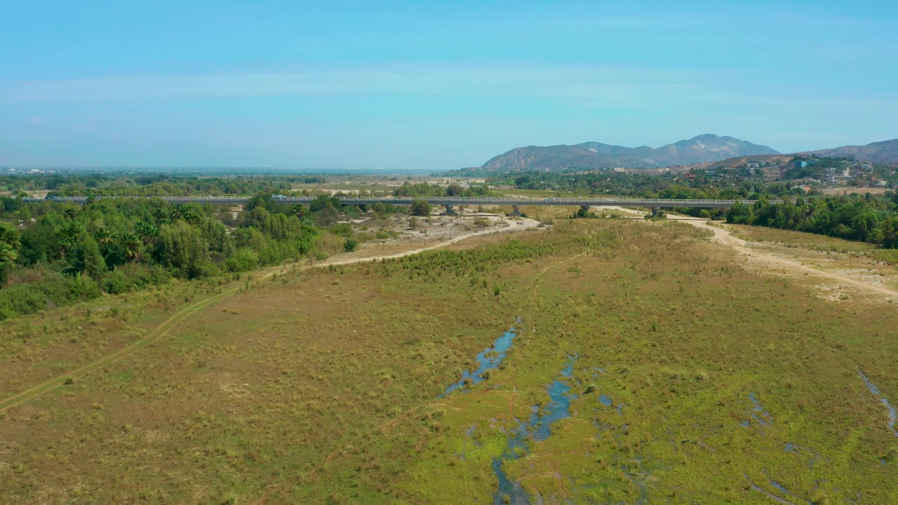 Mid-clip aerial over San Jose Del Cabo with a broad open field, wet patches near the lower frame, a pale dirt road beyond the field, trees, mountains, and no visible people.