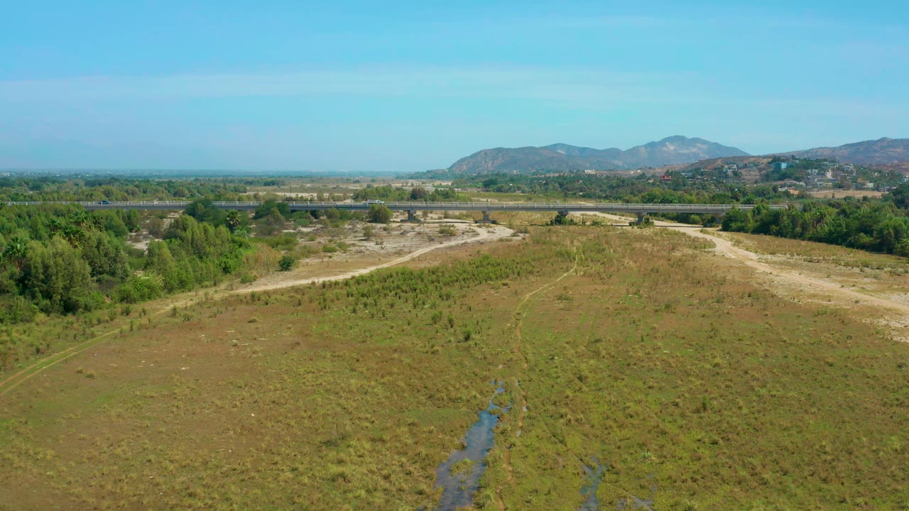Wide aerial landscape with brushy ground, a tree line, highway traffic, and mountains.