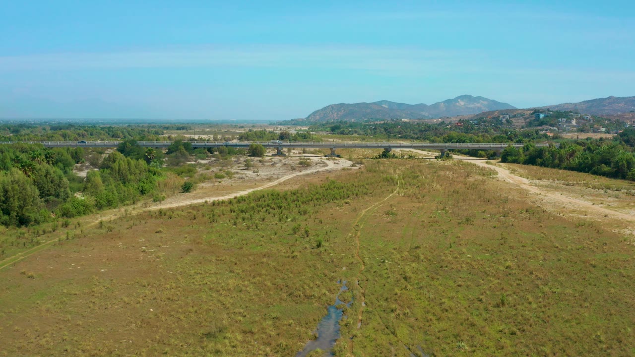San Jose Del Cabo aerial frame showing arid field texture, scattered green shrubs, a road corridor behind a darker tree belt, hazy low mountains, blue sky, and no visible people.
