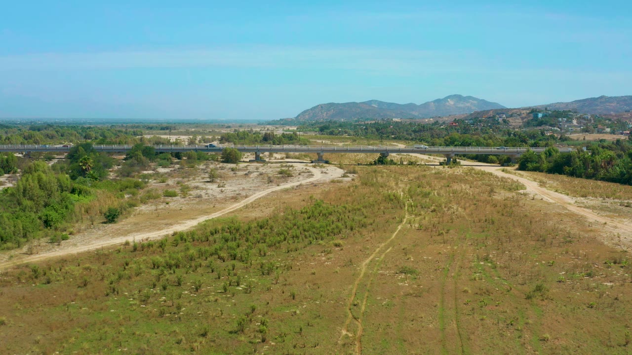 Wide aerial view of bushy dry field, pale road, tree line, and mountains.