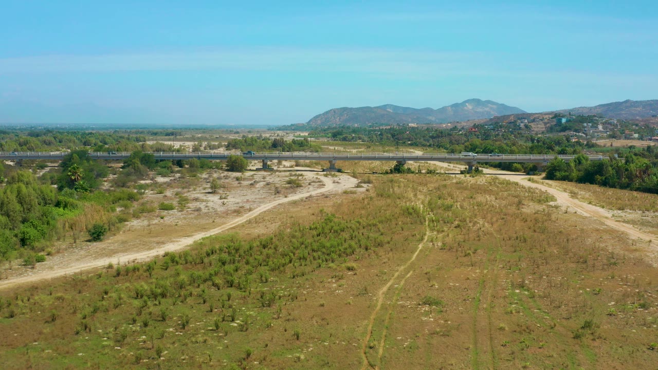Wide elevated view near San Jose Del Cabo showing open brown scrub, faint dirt paths in the foreground, a horizontal road across the midground, green trees, distant low mountains, and no visible people.