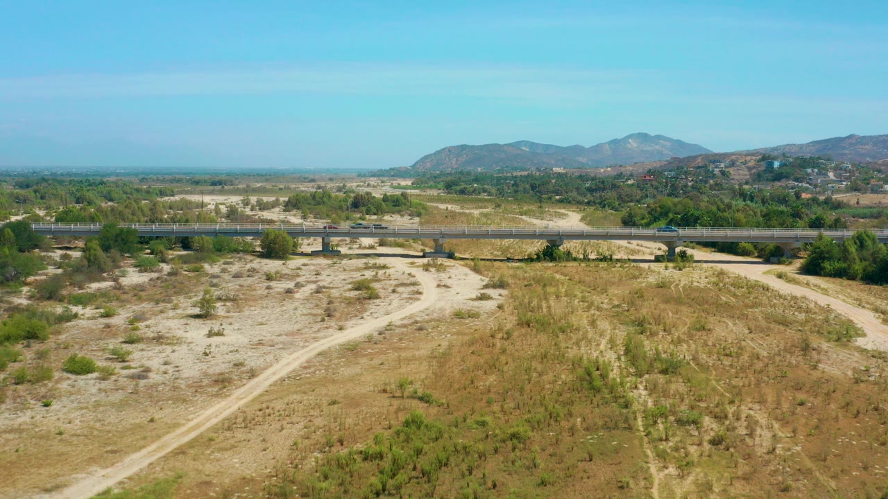 Opening aerial frame over San Jose Del Cabo, Mexico, showing a long bridge with cars crossing a dry sandy wash, dirt tracks, scrub, distant hills, blue sky, and no visible people.