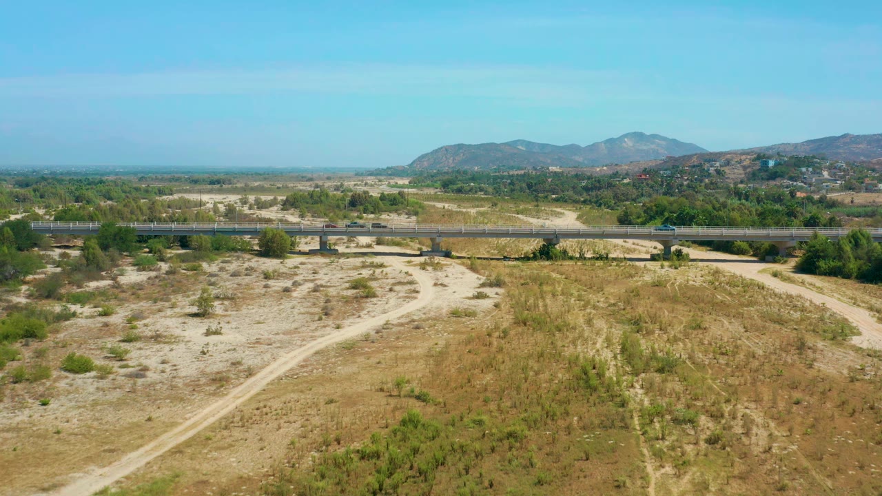 Aerial frame over San Jose Del Cabo, Mexico, with the bridge stretching across the middle of the view, cars spaced along the roadway, pale dirt paths below, green vegetation beyond, and no visible people.