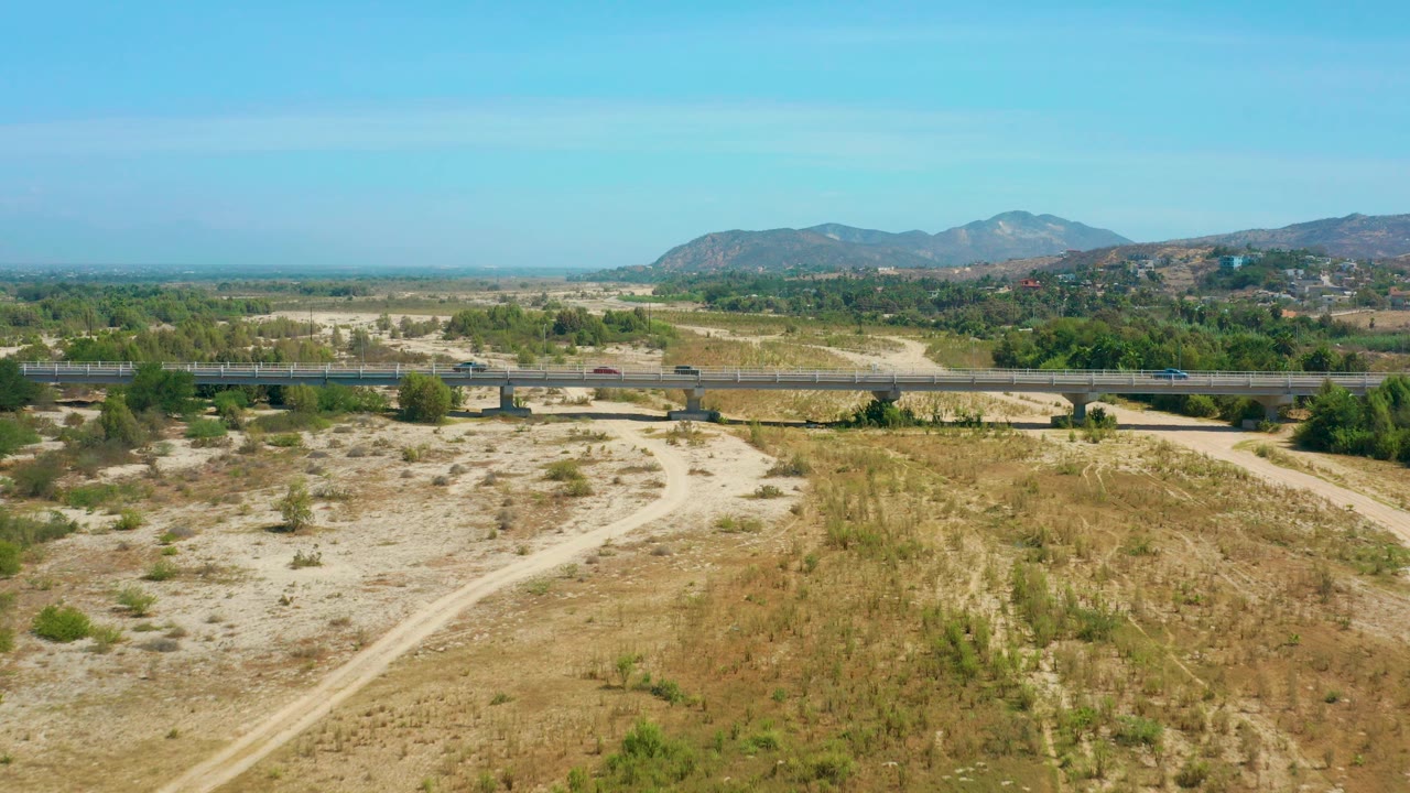 Aerial view in San Jose Del Cabo, Mexico, showing a curved dirt track in the dry foreground, sparse shrubs, cars on the bridge at mid-frame, a green corridor behind it, and no visible people.