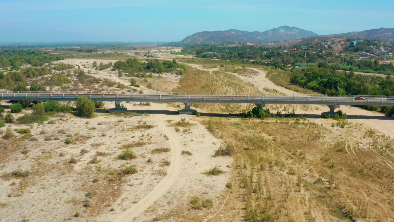 Aerial view in San Jose Del Cabo, Mexico, showing a concrete bridge crossing a sandy wash with green trees, dry tracks, low hills, blue sky, and no visible people.