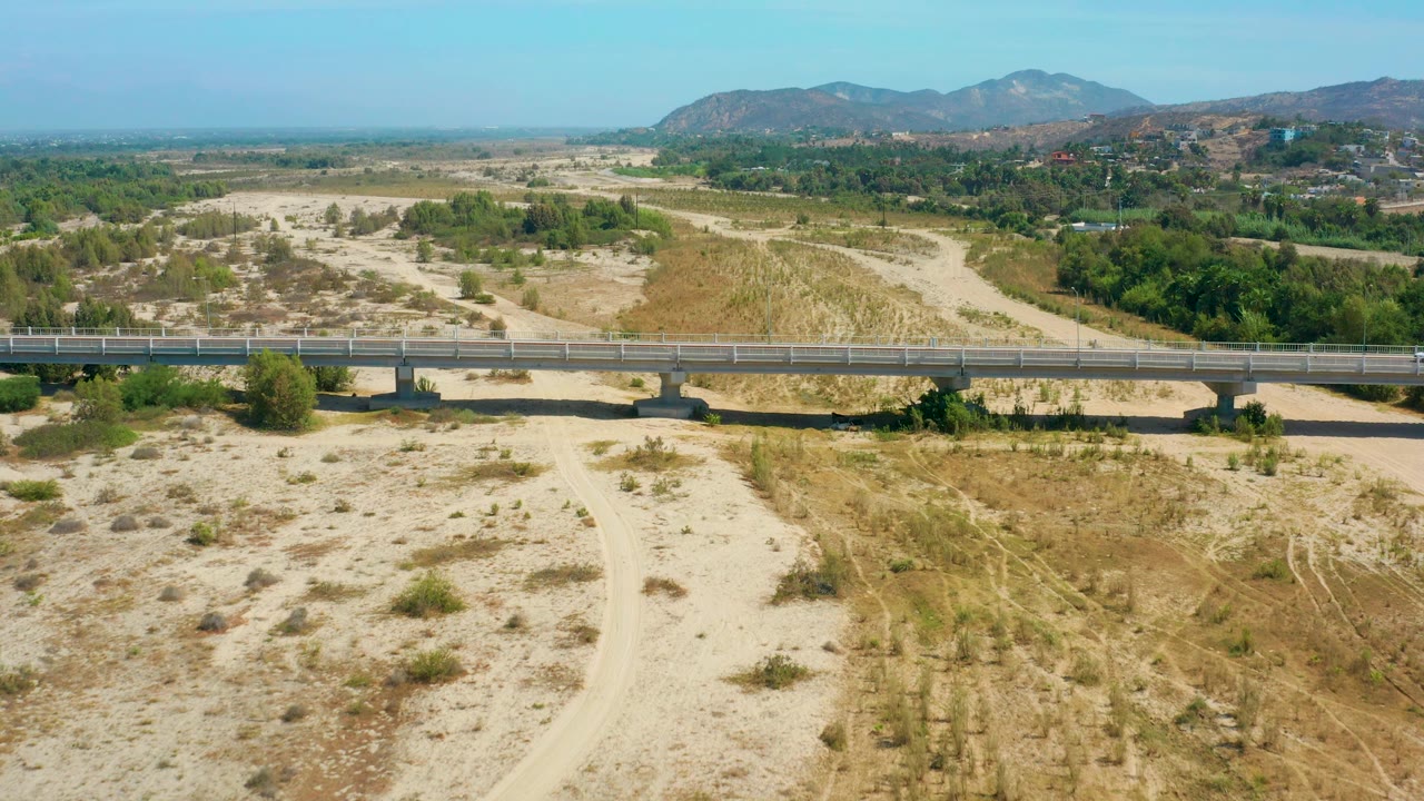 Aerial San Jose Del Cabo frame with a white car on the concrete bridge, sandy wash and dirt road below, green trees at the edge, hills in the background, and no visible people.