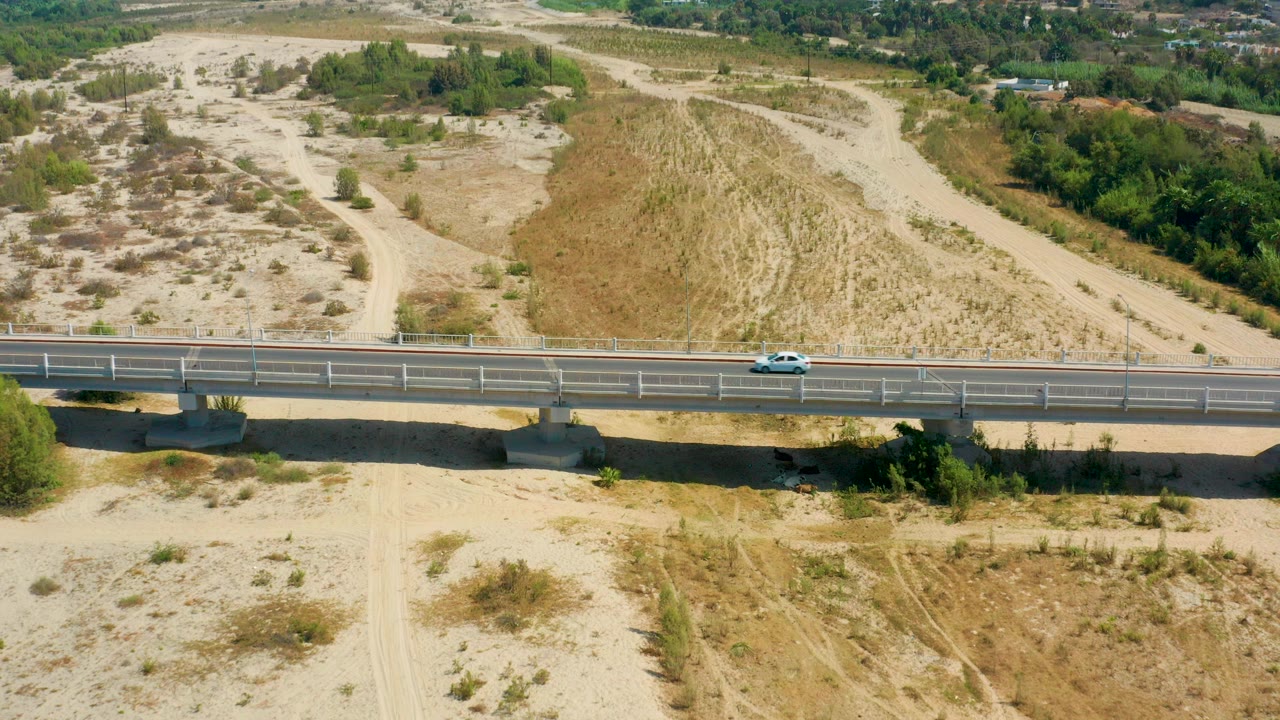 A white car travels along a paved bridge in San Jose Del Cabo, Mexico, with sandy scrubland, dirt tracks, green vegetation, and no visible people.