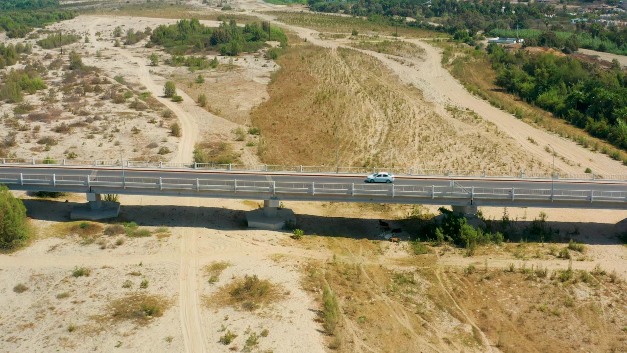 The white car continues across the bridge in San Jose Del Cabo, Mexico, passing below sandy hills, dirt paths, scrub patches, and distant greenery with no visible people.