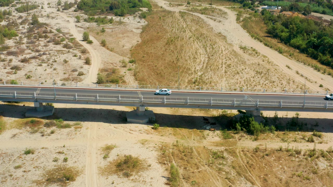 A small white car moves along the bridge in San Jose Del Cabo, Mexico, with a pale dirt road climbing through the dry hillside behind it and no visible people.