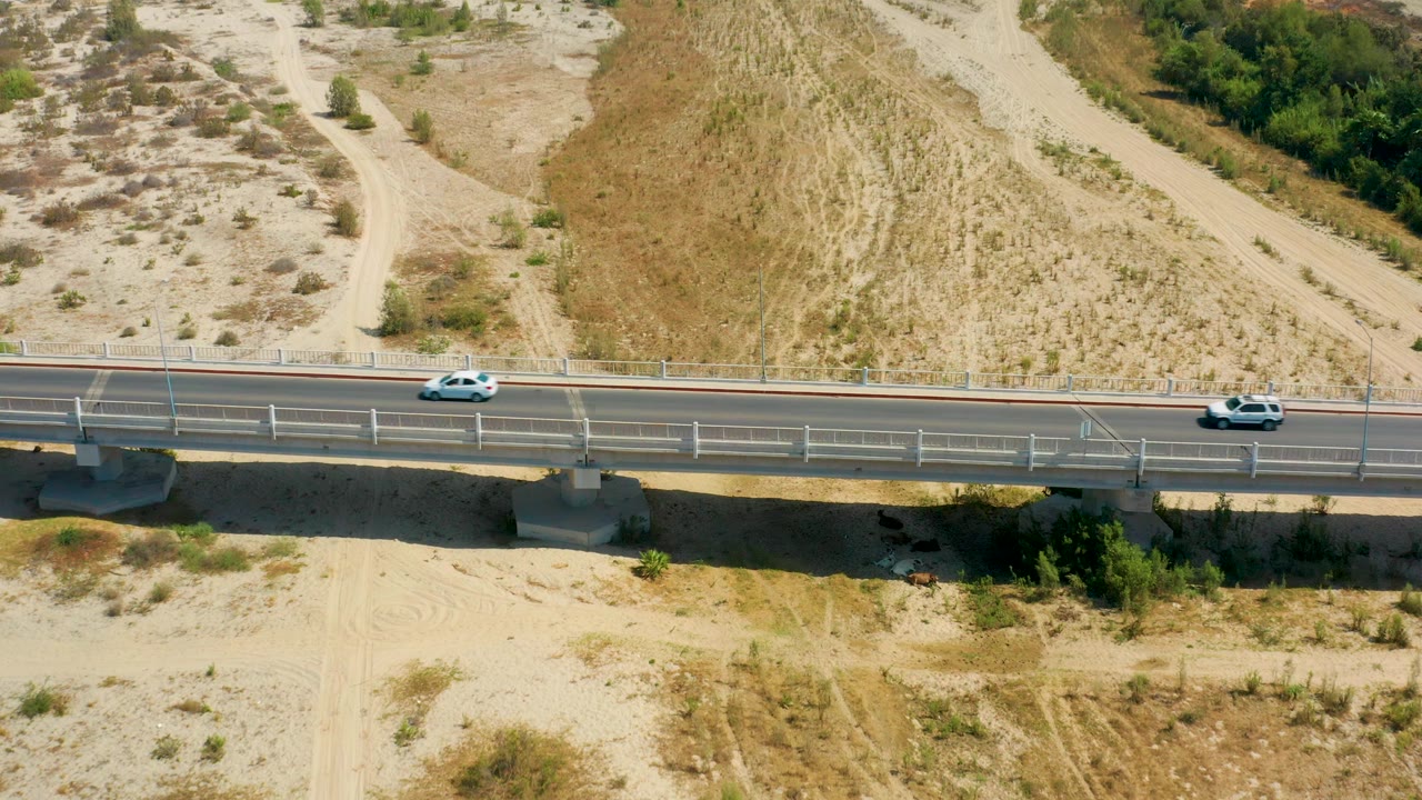 A white car and a dark car appear on the bridge in San Jose Del Cabo, Mexico, separated across the lanes above sandy ground, bridge pillars, and no visible people.