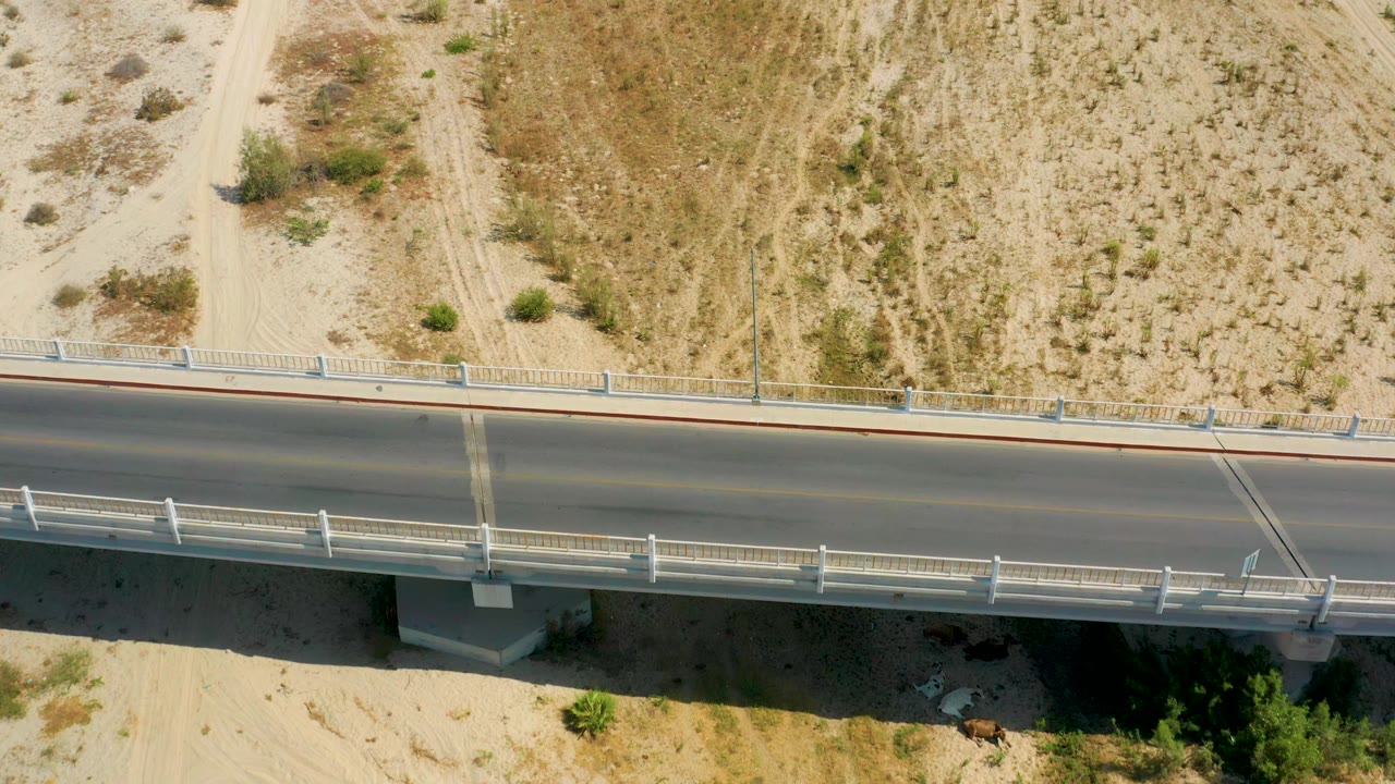 Aerial top-down frame in San Jose Del Cabo, Mexico with the paved road spanning the image, pale dirt tracks above it, bridge railings, and no visible people.