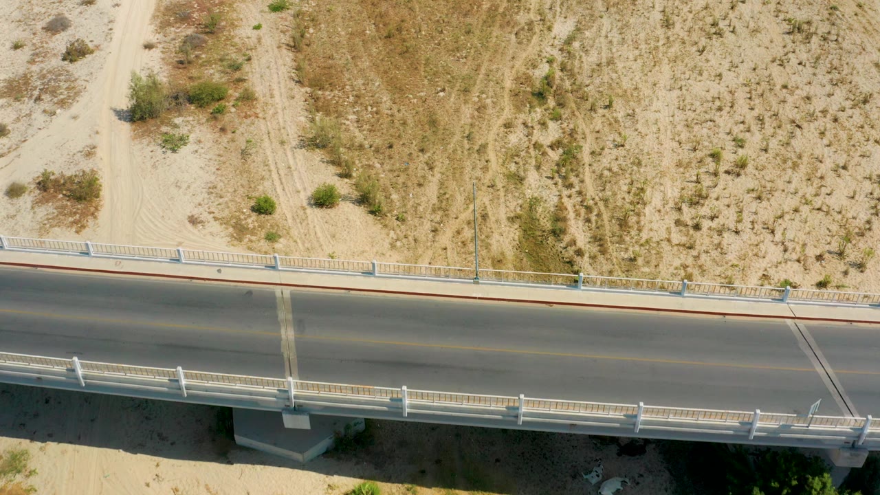 Top-down San Jose Del Cabo, Mexico frame showing the bridge-like road, white side barriers, sandy shoulder, scrub vegetation, and no visible people.