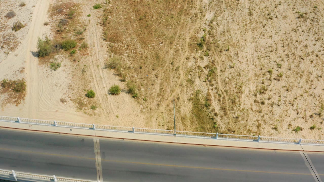 Overhead frame in San Jose Del Cabo, Mexico where the road drops toward the lower edge, leaving sandy tracks, dry brush, and no visible people across most of the view.