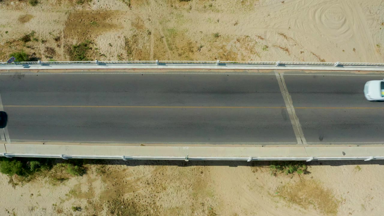 Overhead view of a concrete road in San Jose del Cabo with a light vehicle partly entering at the left edge, sandy shoulders, scrub, guardrails, and no visible people.