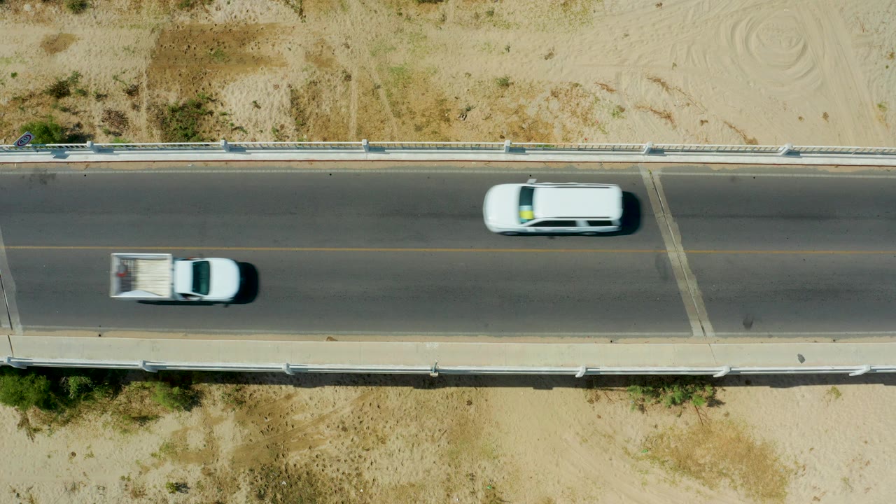 A white car and a white pickup truck appear on the concrete road in San Jose del Cabo, separated along the lane with sandy shoulders and no visible people.