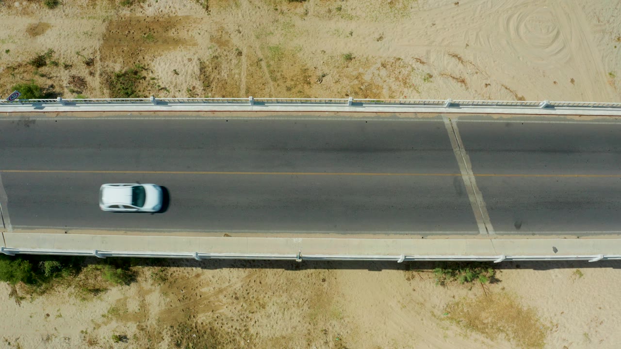 Overhead San Jose Del Cabo road view with the white car nearer the middle of the bridge, pale sand above and below the road, and no visible people.
