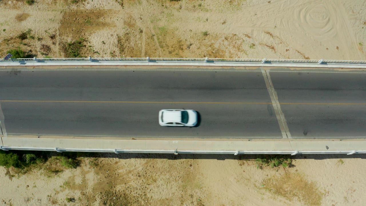 A white car continues along the paved bridge in San Jose Del Cabo, Mexico, with desert sand, guardrails, and faint spiral marks visible nearby, with no visible people.