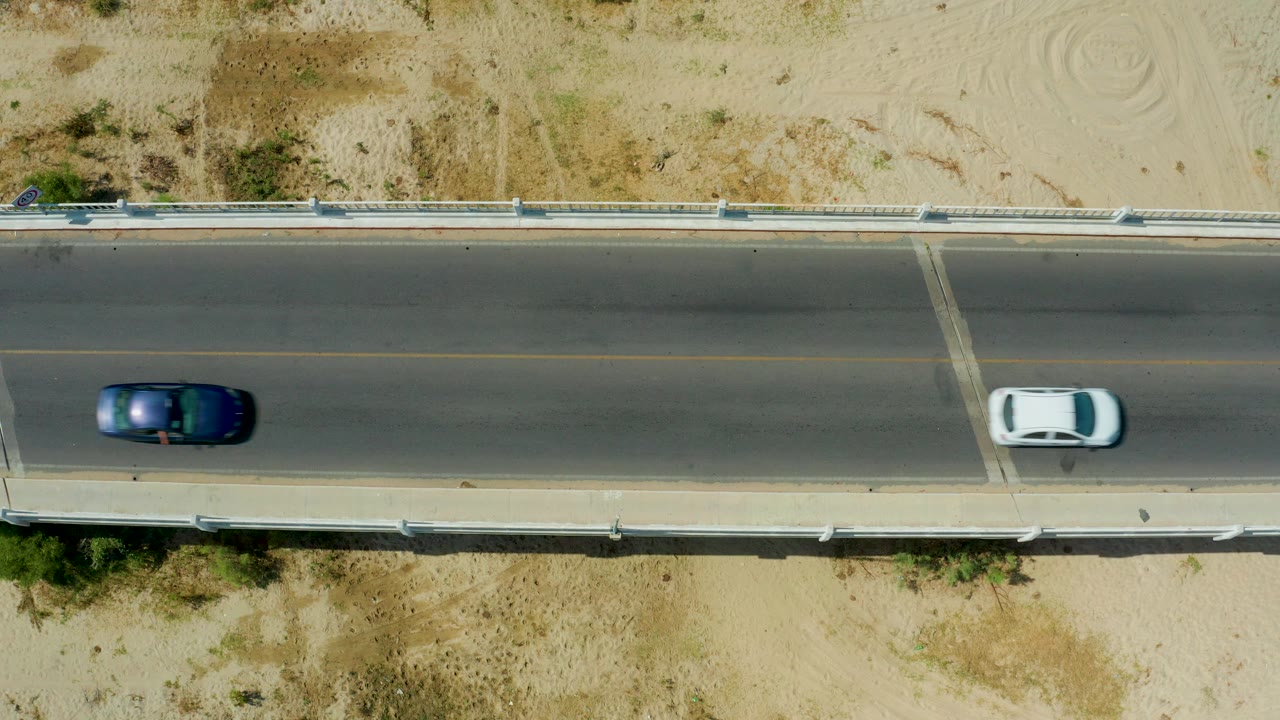 Overhead view of a dark car entering the bridge scene in San Jose Del Cabo, Mexico, with a white car farther along the road and no visible people.