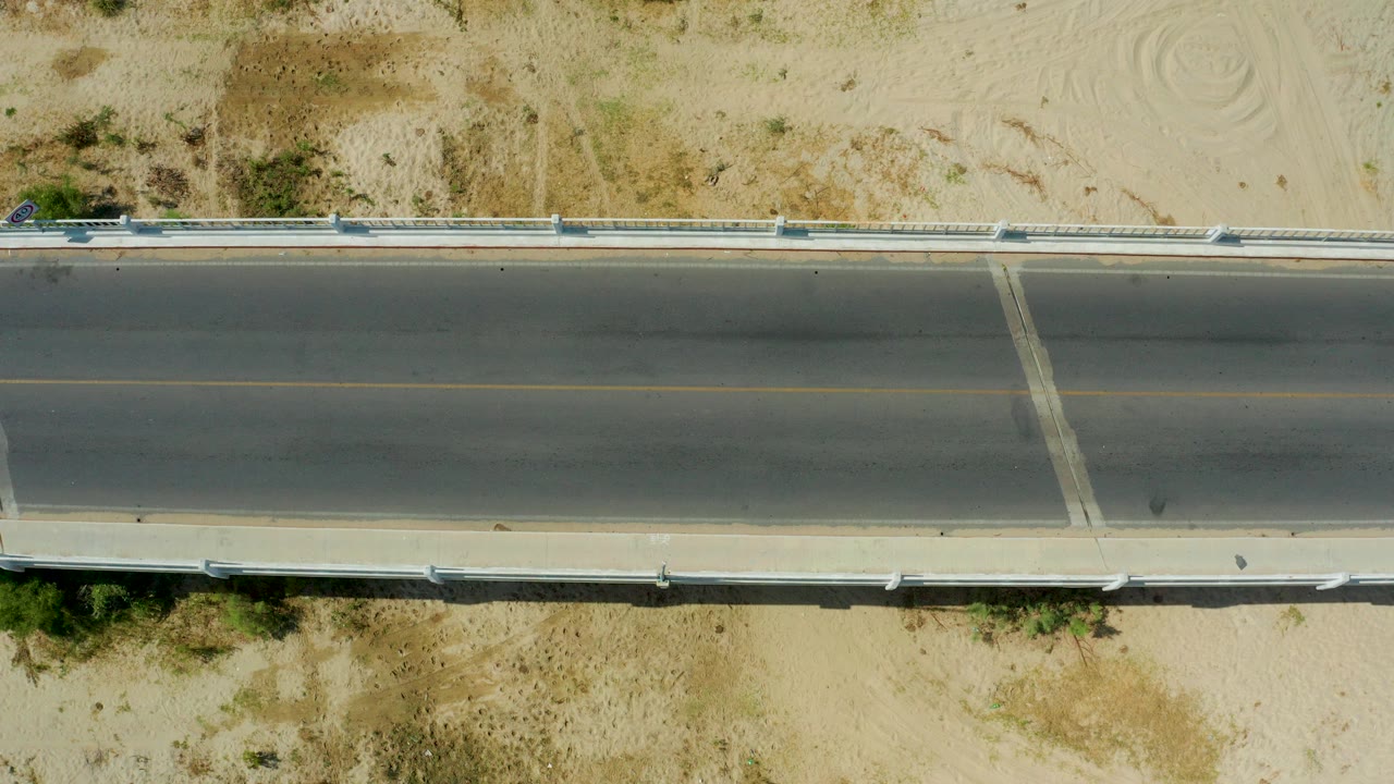 Top-down view of a two-lane road in San Jose Del Cabo, Mexico, bordered by pale sand, scrub, guardrails, and a small car near the upper left edge, with no visible people.