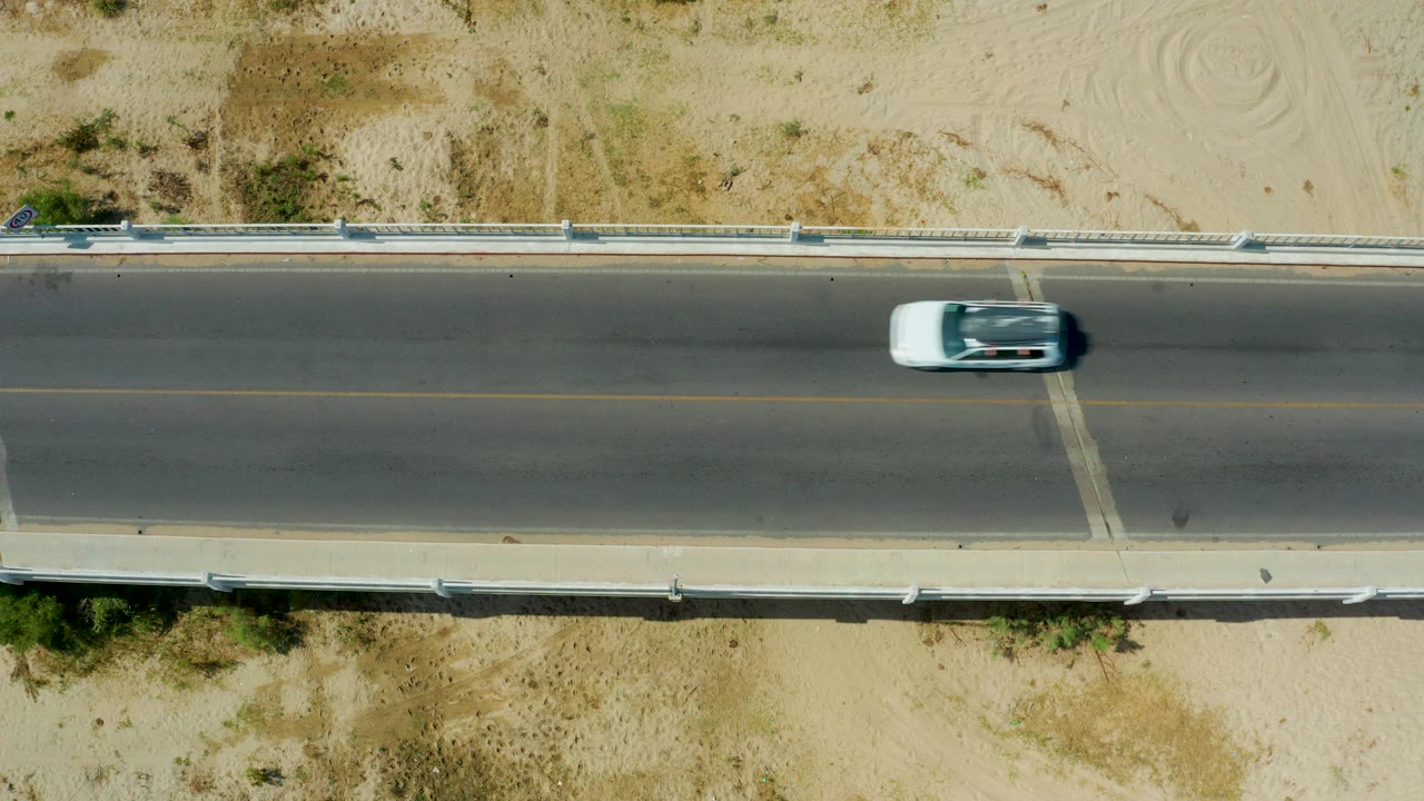 A light-colored car appears near the right side of the two-lane road in San Jose Del Cabo, Mexico, with sandy ground and guardrails around it and no visible people.