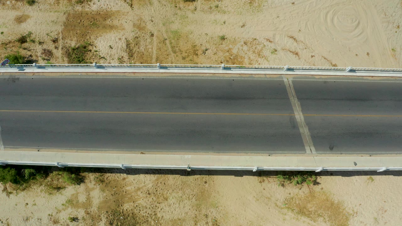 A straight blacktop road spans the frame in San Jose Del Cabo, Mexico, bordered by pale sand, scrub patches, and metal guardrails, with no visible people.