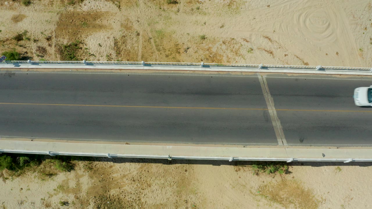 Top-down view of a paved road in San Jose Del Cabo, Mexico, bordered by sandy ground and guardrails, with a white car entering near the right edge and no visible people.