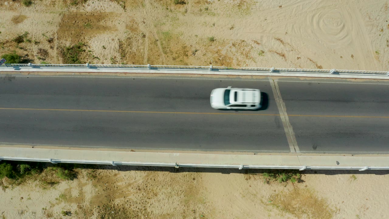 A white SUV passes through the right side of the overhead road frame in San Jose Del Cabo, Mexico, with sandy ground and guardrails visible and no visible people.