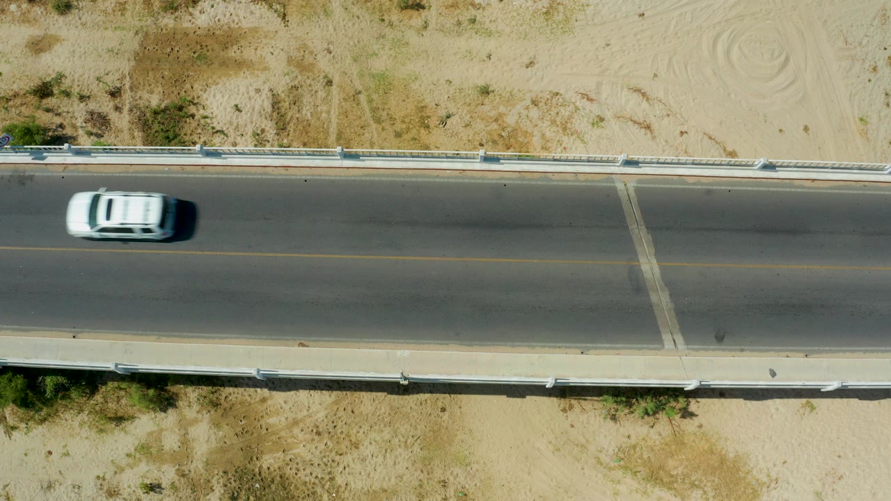 The white vehicle is partly exiting the left edge of the overhead San Jose Del Cabo road view, leaving open pavement, sandy shoulders, and no visible people in the frame.