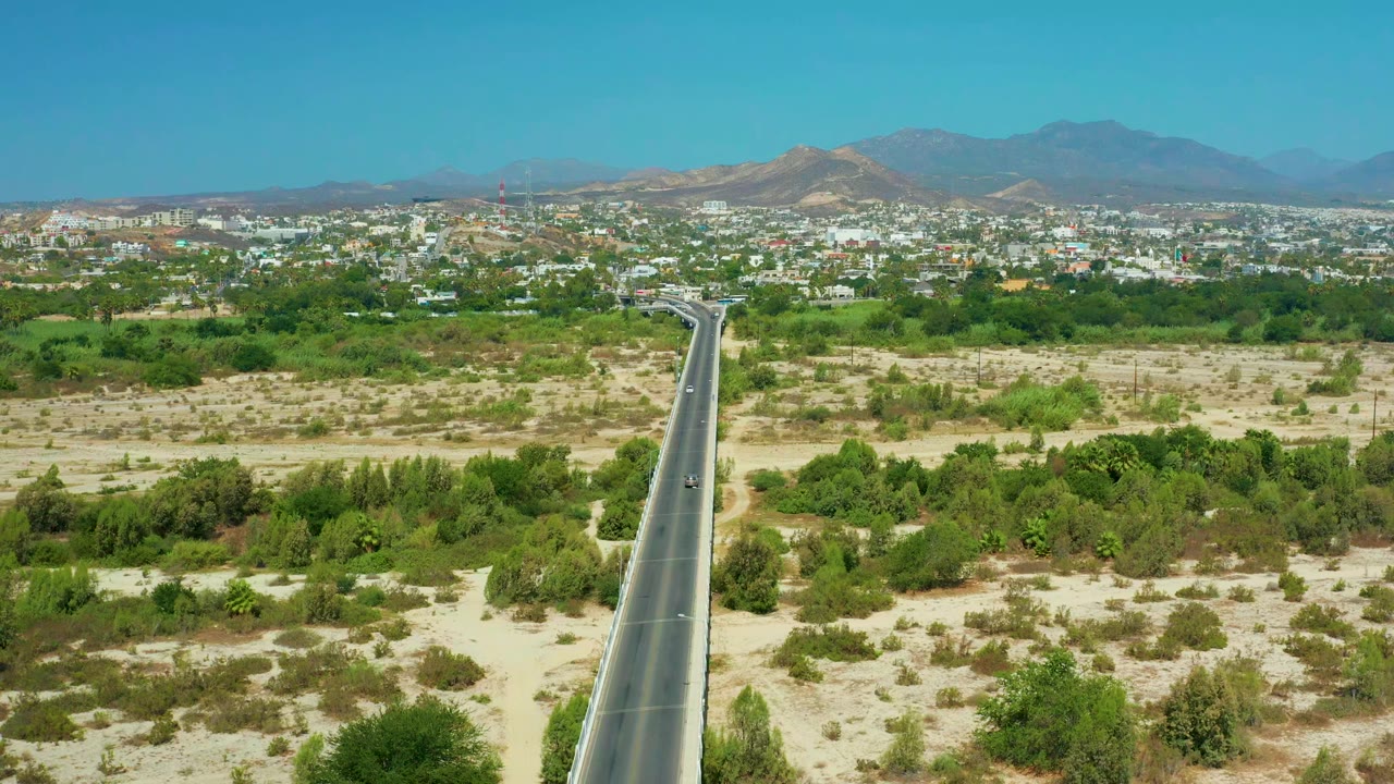 Aerial view in San Jose del Cabo, Mexico, showing a straight road through pale scrub and green trees toward a low city and hazy mountains, with no visible people.