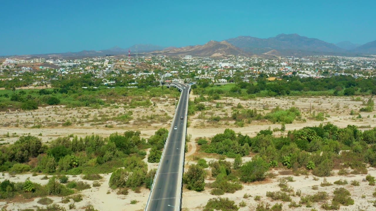 Aerial view in San Jose del Cabo with a paved road entering from the lower left, green scrubland and trees across sandy ground, a city band in the distance, mountains beyond it, and no visible people.