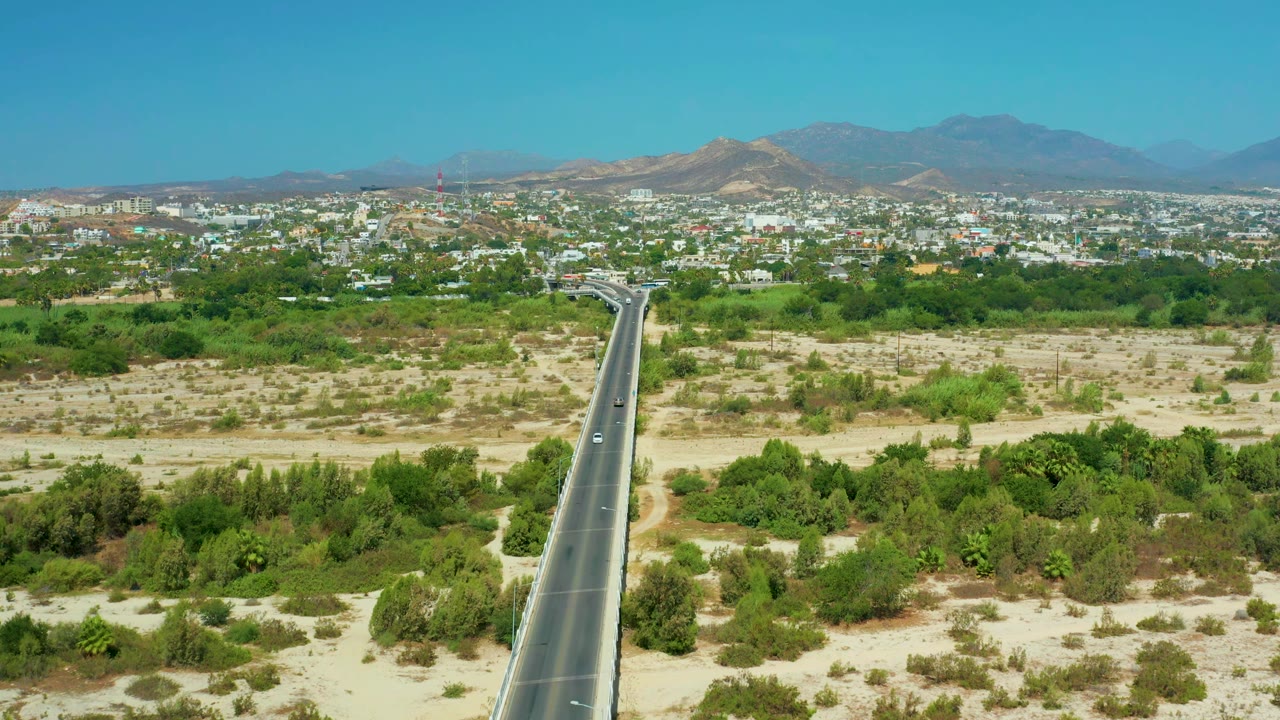 Aerial road view with sandy open land, dense green trees, city buildings, and mountains.