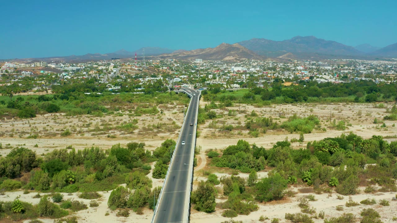 High aerial view in San Jose del Cabo with a straight paved road on the right side, a small white vehicle on the road, sandy scrubland and green trees below, distant city blocks, and no visible people.