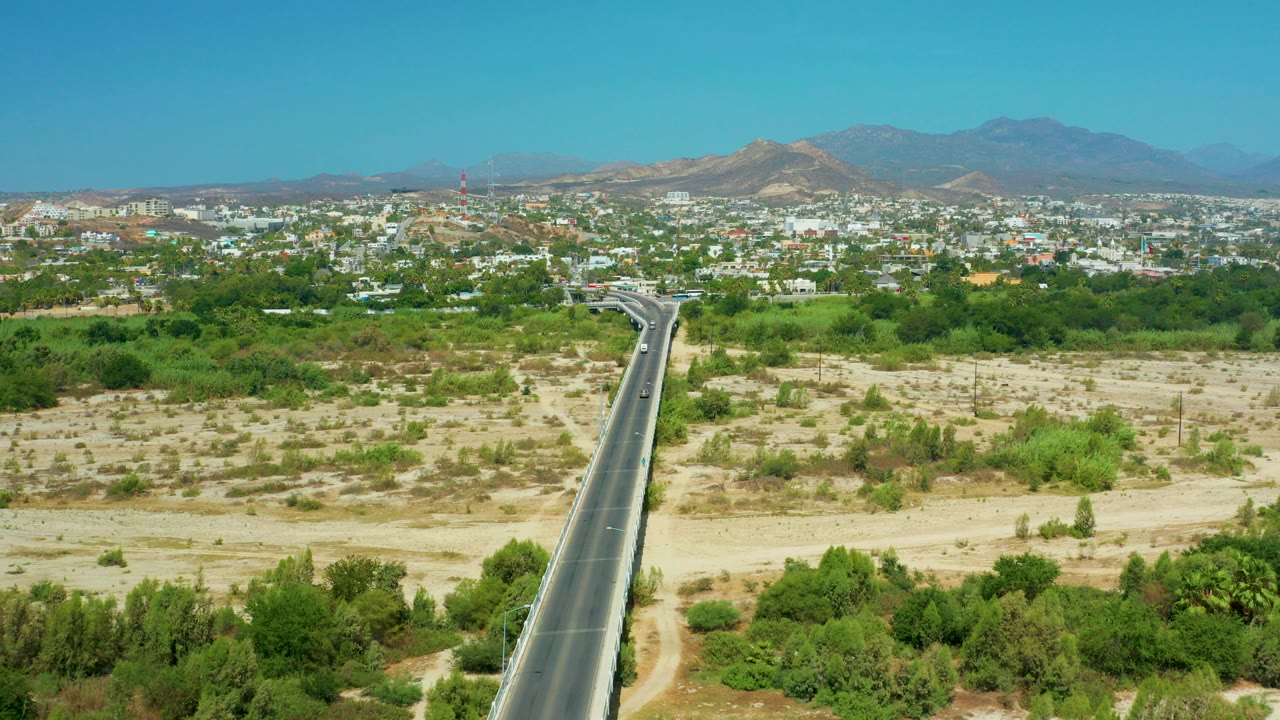 Aerial view in San Jose Del Cabo, Mexico, with a straight highway entering from the lower left, dry scrubland and green trees around it, town buildings in the distance, mountains beyond, and no visible people.