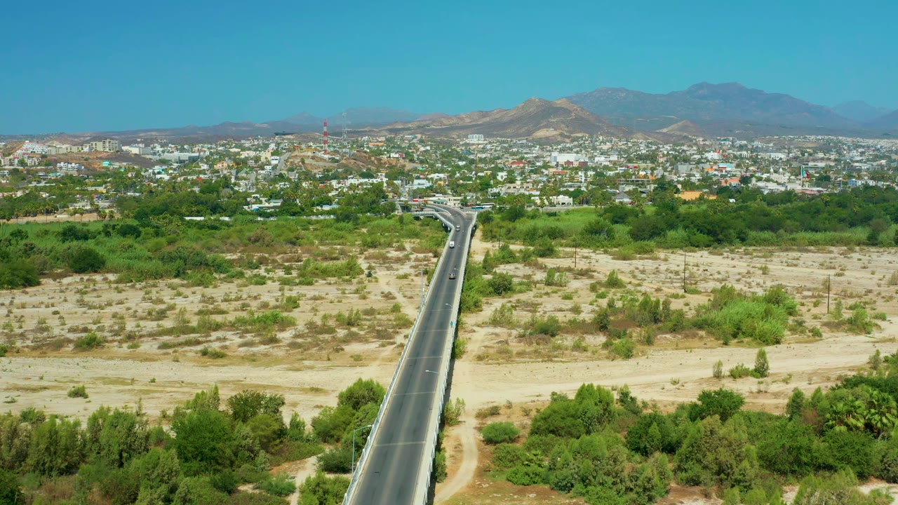 Aerial frame of San Jose Del Cabo, Mexico, showing the road slightly more centered through sandy open ground, with a few vehicles on the pavement, dense green vegetation near town, distant buildings, mountains, and no visible people.