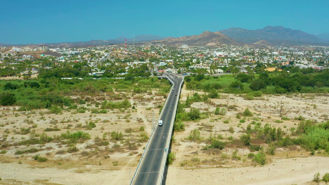 Aerial frame in San Jose Del Cabo, Mexico, with sparse cars on the paved road, green brush across sandy ground, a spread of white buildings, distant mountains, and no visible people.