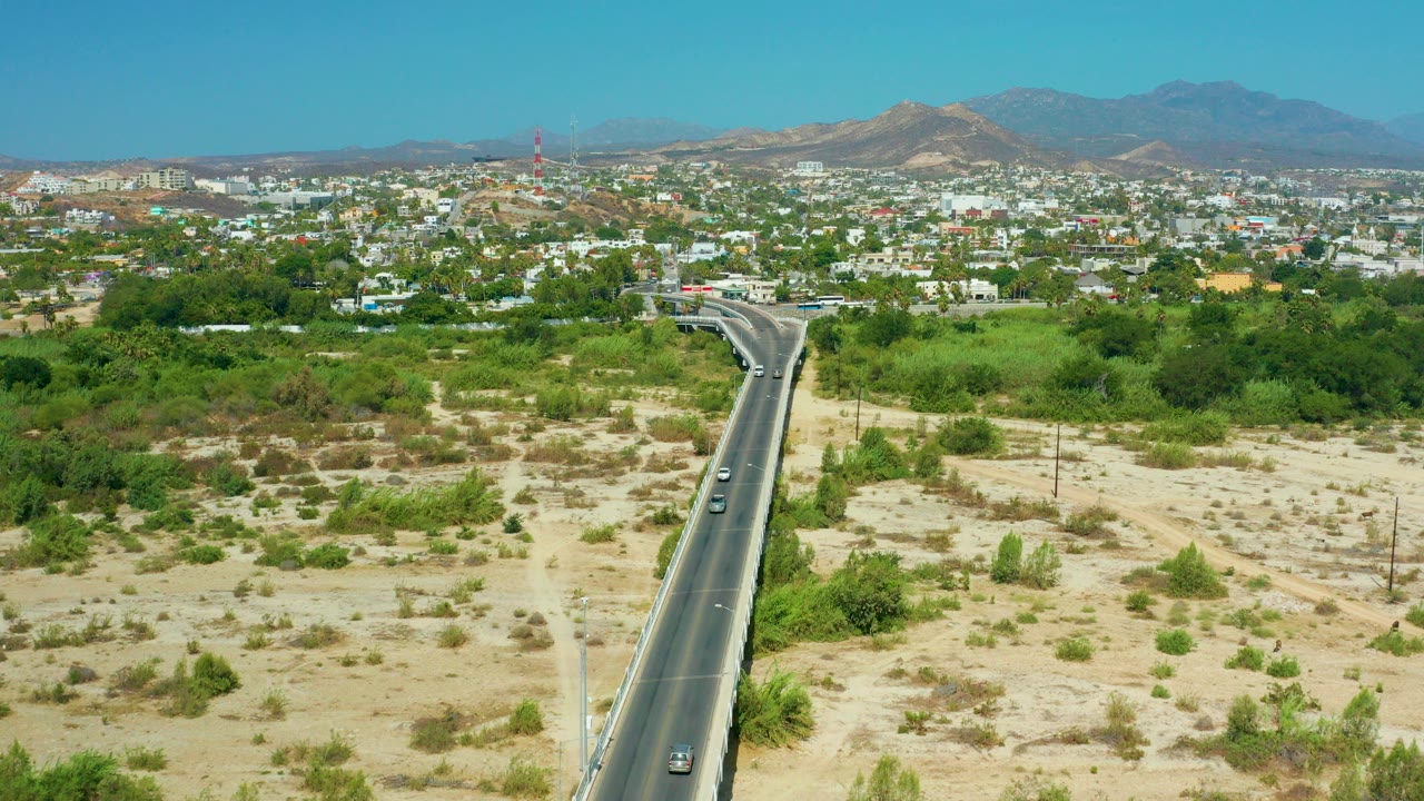 Opening aerial frame of San Jose Del Cabo showing a paved road running through sandy scrub and green trees toward low buildings, distant hills, blue sky, and no visible people.
