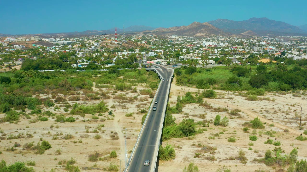 Drone view of cars on a road leading toward low buildings and mountains.