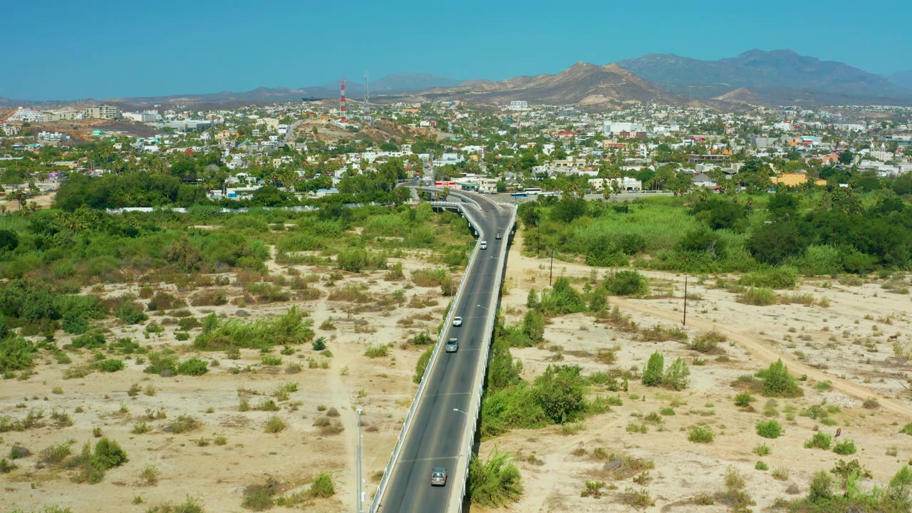 Third frame shows the paved road entering from the lower edge, surrounded by pale sandy ground and green brush, with San Jose Del Cabo buildings and hazy mountains across the background, and no visible people.
