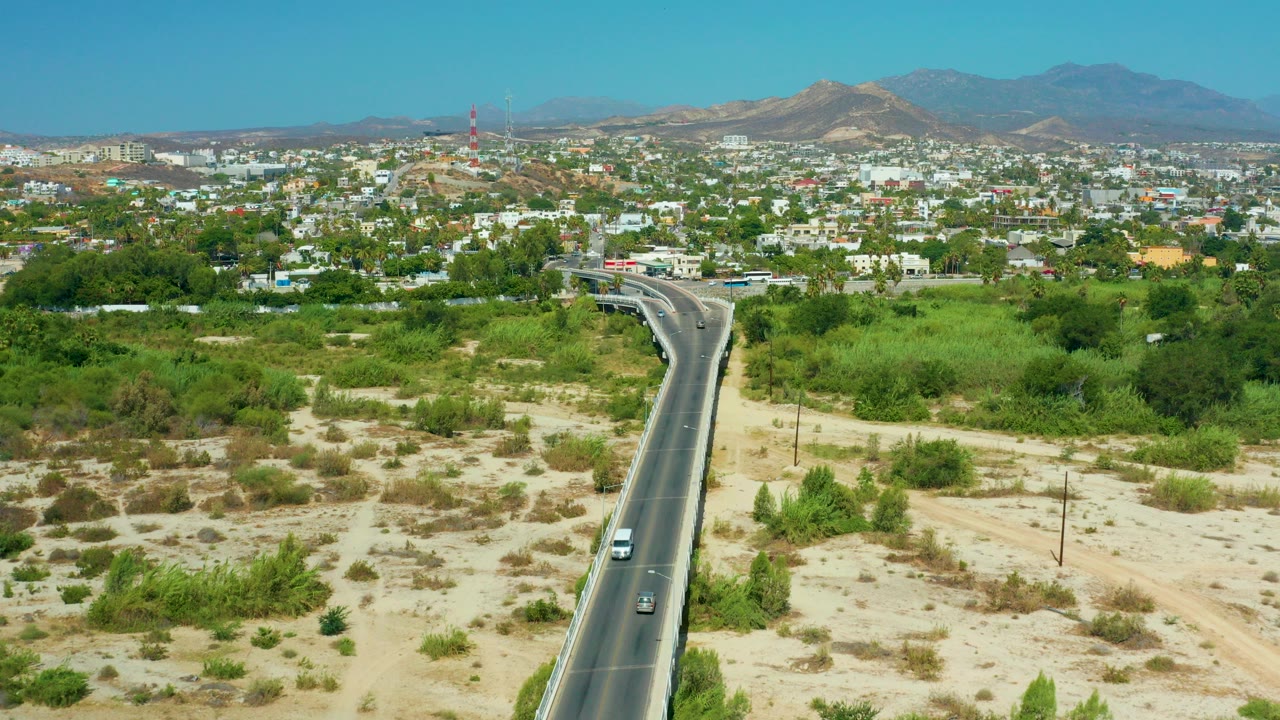 Elevated view in San Jose del Cabo, Mexico, with a black road entering from the lower left, sandy scrubland on both sides, low buildings, green trees, distant hills, and no recognizable people.