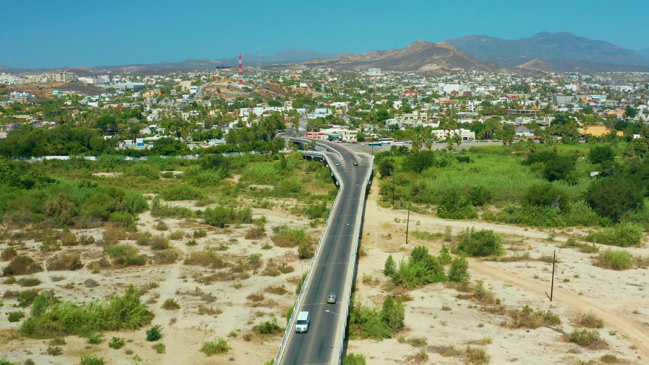 Aerial view in San Jose del Cabo, Mexico, with the paved road cutting through pale scrubland toward dense green trees, low buildings, a red-and-white tower, mountains, and no recognizable people.