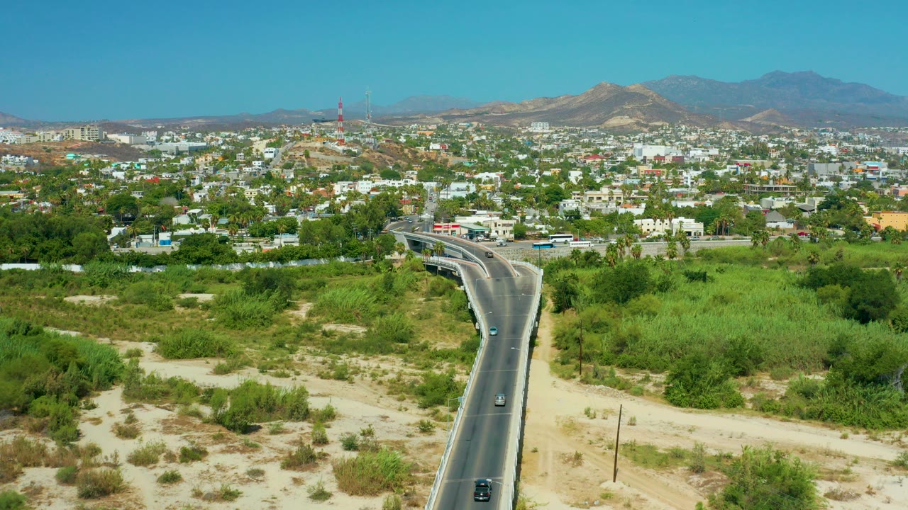 Aerial view over San Jose Del Cabo with a curving road in the foreground, green vegetation, low city buildings, distant mountains, and no visible people.