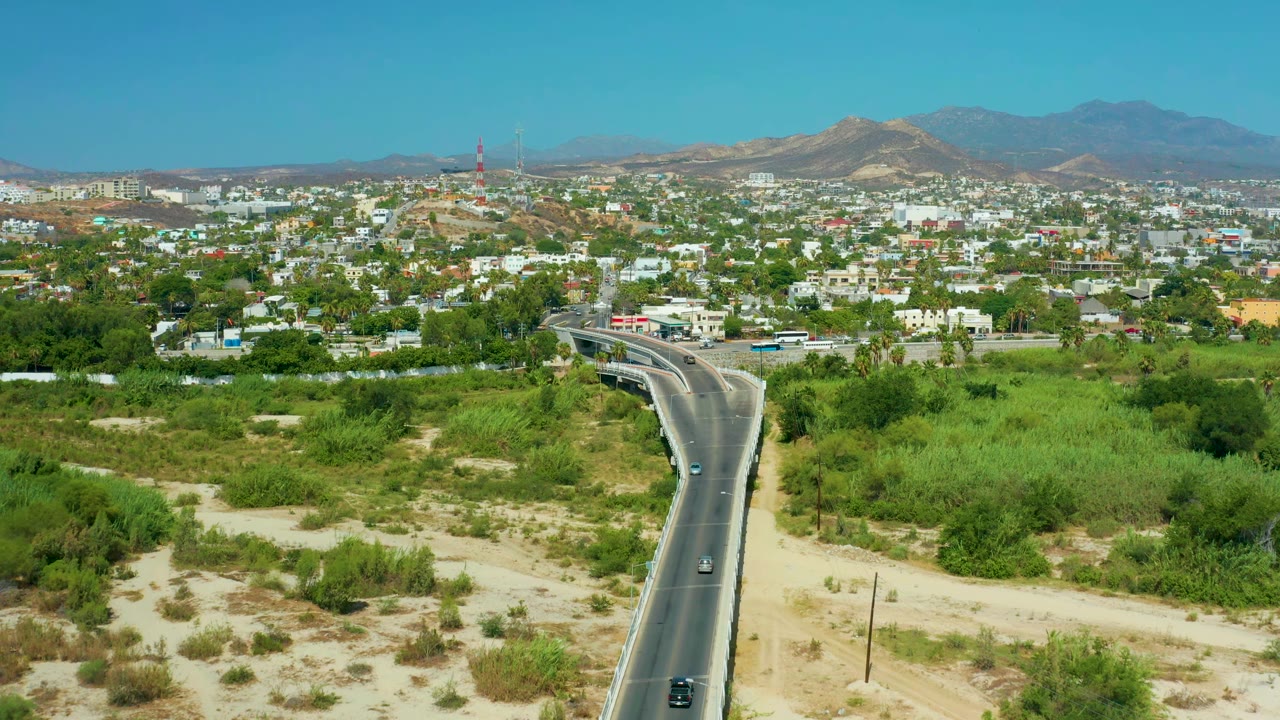 Elevated San Jose Del Cabo view with small cars on the road, dense green brush on both sides, white city buildings, hills, mountains, and no visible people.