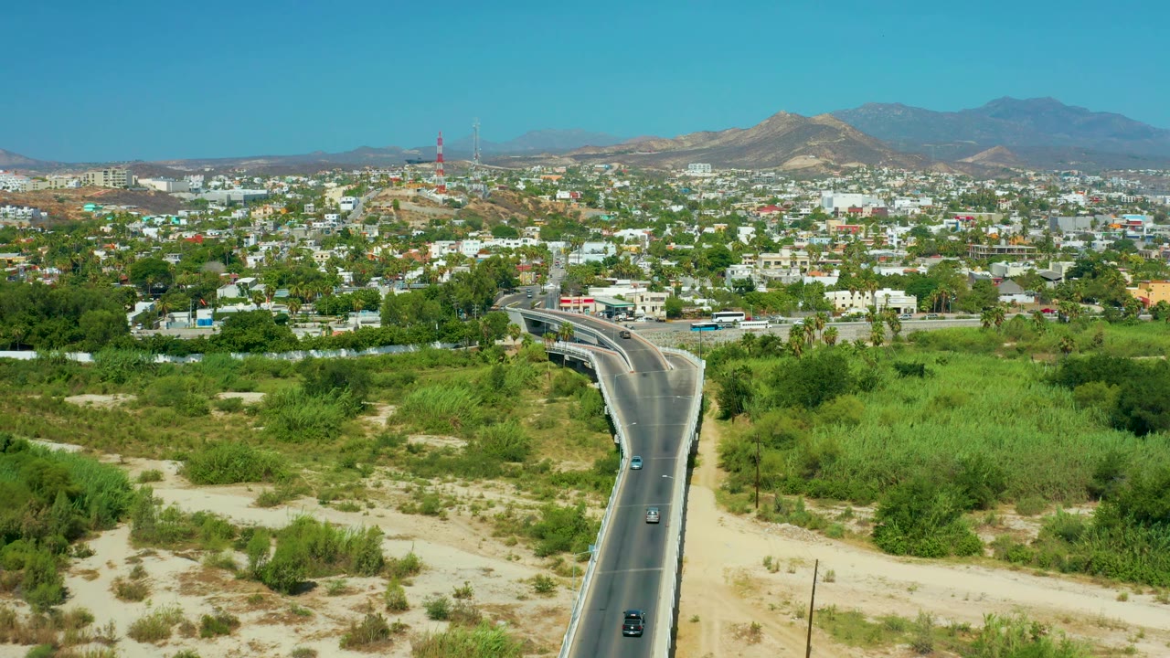 Wide San Jose Del Cabo aerial showing a paved road rising from the lower frame, scattered cars, green vegetation, a low city skyline, brown hills, mountains, and no visible people.