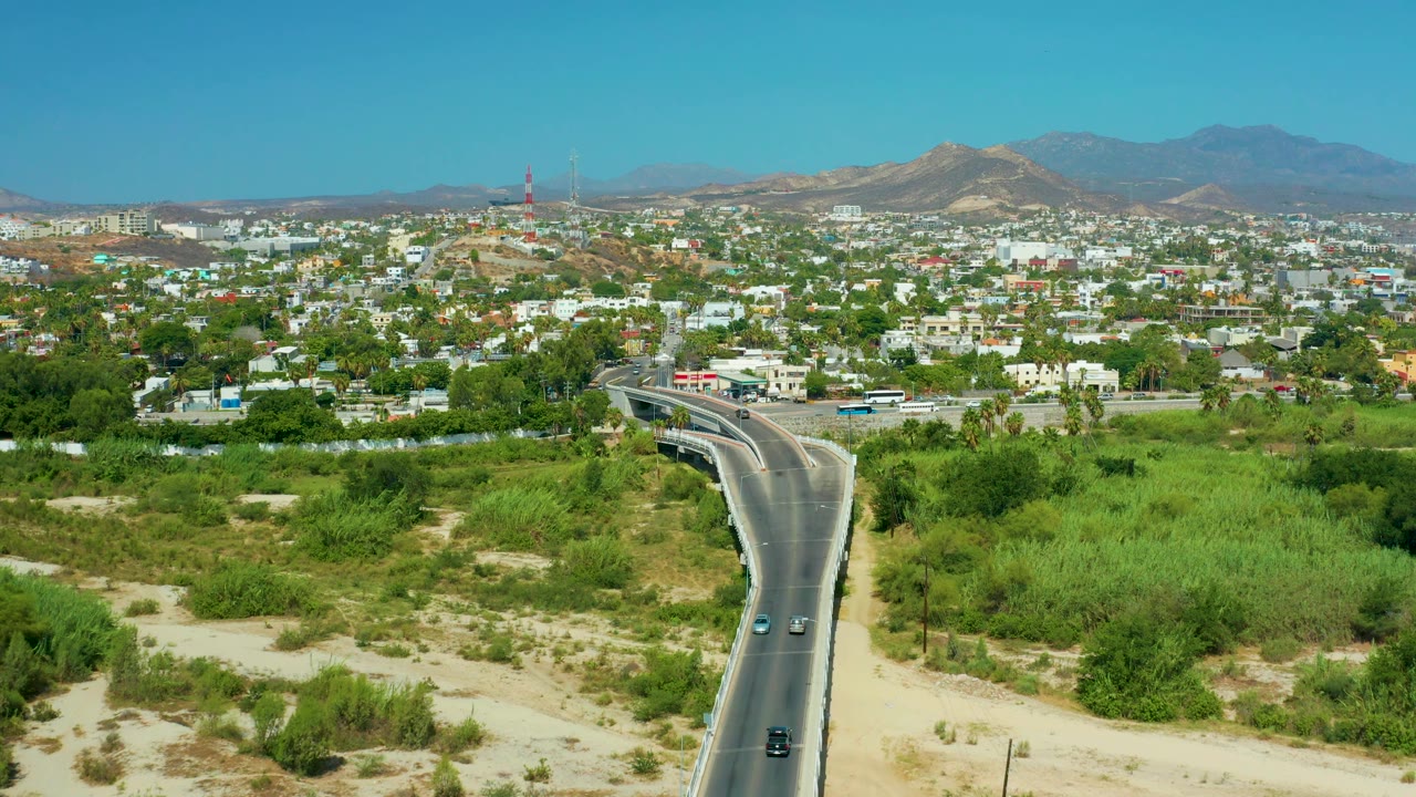 San Jose Del Cabo aerial frame with the road bending through a green corridor, a spread of low buildings ahead, distant towers, mountain ridges, and no visible people.