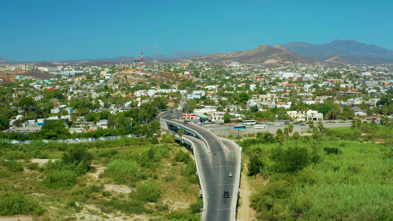 San Jose Del Cabo aerial frame showing the road centered at the bottom, vehicles on the pavement, dense green areas, low urban buildings, distant mountains, and no visible people.